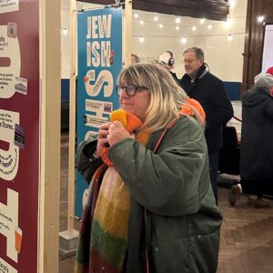 A woman wearing glasses is clenching an orange hat in her hands, she is surrounded by posters of famous books.