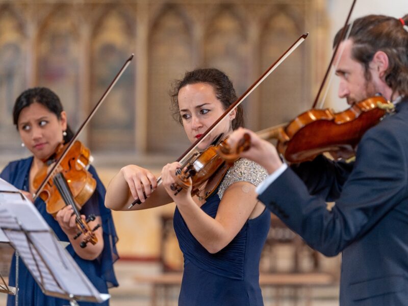 Three violinists, two women and a man, are performing while looking at their music sheets at the King's Lynn Festival.