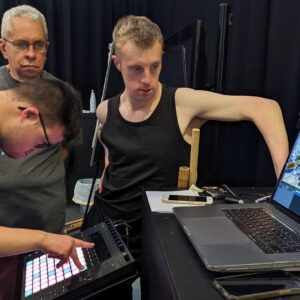 Three men surrounding a laptop playing a video on video editing software. One of the men is holding his arm up at an angle.