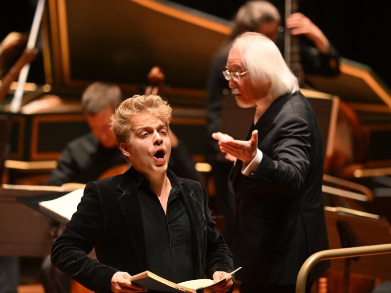 An orchestral performance with an older man with shoulder-length white hair conducting the orchestra while a singer is in the forefront with his mouth open singing and holding a book.