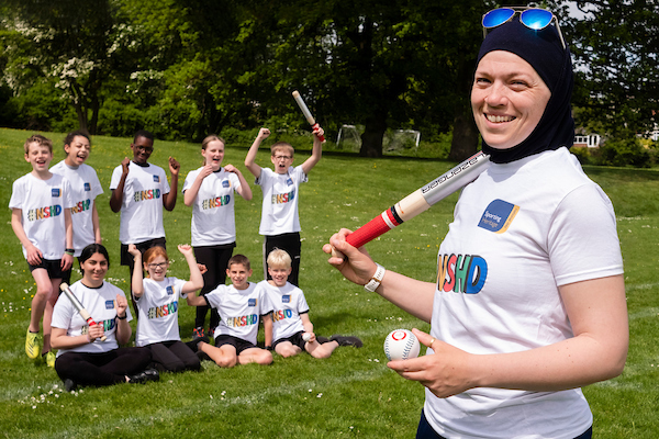 A woman in a black hijab stands with a baseball bat in her hands, there is a group of children behind her also holding baseball bats.