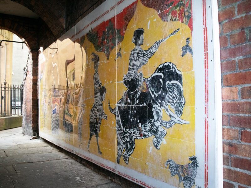 The artwork, the path to happiness as a mural on a wall in Wakefield. There are several people on a yellow background. Creating a sort of tapestry.