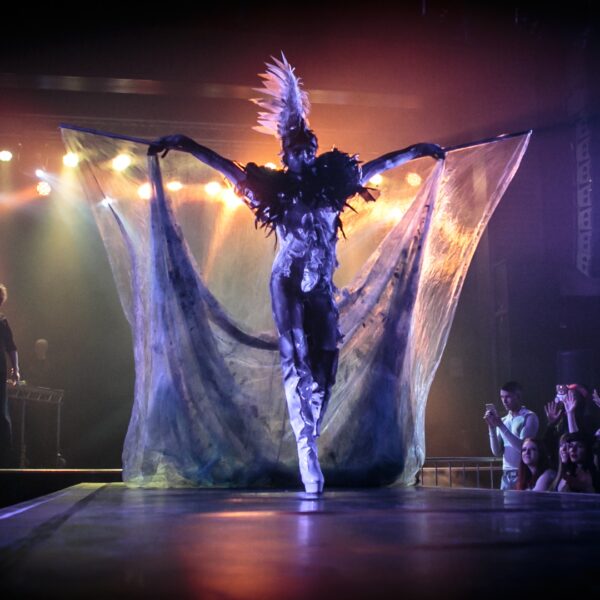 A person on a runway with their arms outstretched, covered in yellow, orange and purple lights. They wear high white boots and a white headdress. Surrounding the runway are cheering people.